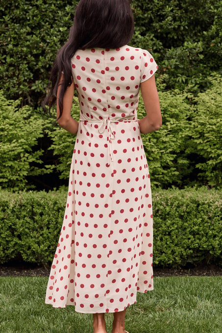 A woman with long dark hair stands barefoot outdoors among green bushes, wearing the Ivy City Co Fiona Dress in Red Polka Dot—an ankle-length, cream dress with red polka dots and a tie waist.