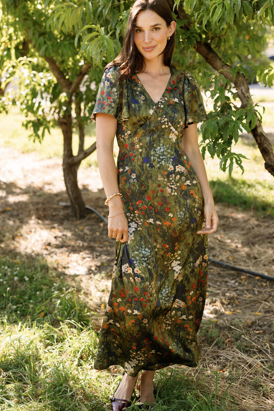 A woman stands outdoors in a grassy area, wearing the Ivy City Co Fiona Flutter-Sleeve Maxi Dress in Impressionist Bloom. Trees with green leaves are in the background, and sunlight filters through the branches.