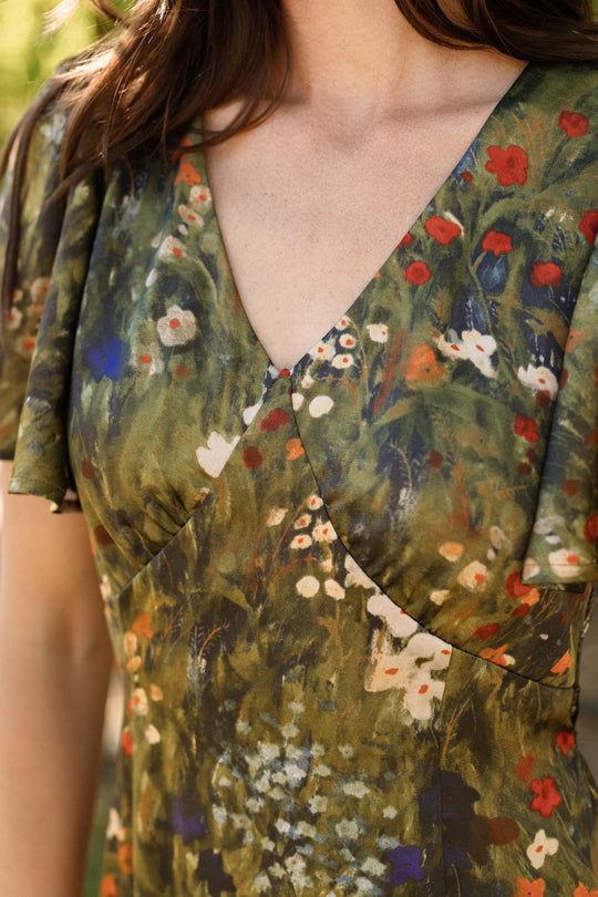 A close-up of a woman wearing the Ivy City Co Fiona Flutter-Sleeve Maxi Dress in Impressionist Bloom—a green satin maxi dress adorned with red, white, and blue floral prints. The dress features a V-neckline and flutter sleeves, with only her torso and lower neck visible.