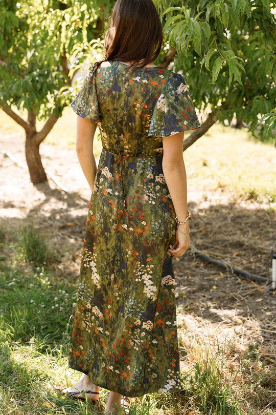 A woman stands outdoors facing away, surrounded by sunlit trees and grass, wearing the Ivy City Co Fiona Flutter-Sleeve Maxi Dress in Impressionist Bloom—a long satin dress adorned with flutter sleeves and a vibrant floral print.