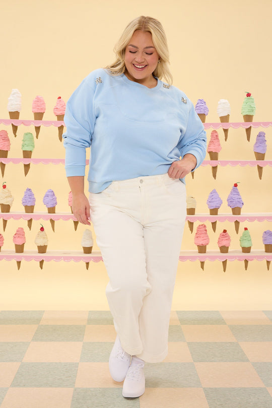A smiling woman stands with one hand in her pocket, wearing the Ivy City Co Casey Crewneck Sweatshirt in Light Blue embellished with subtle crystal details, paired with white pants. Behind her, pastel shelves are adorned with a colorful display of ice cream cones.