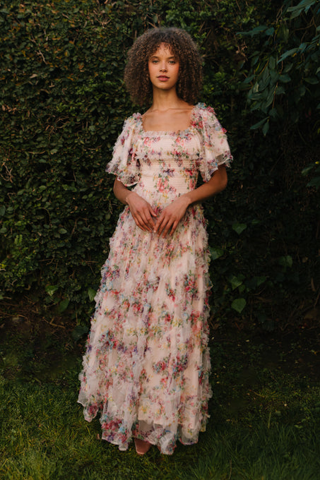 A woman with curly hair stands before lush greenery, calmly looking at the camera while wearing Ivy City Co’s Florentina Dress in Pink Floral—a flowing maternity style with ruffled layers and short sleeves.