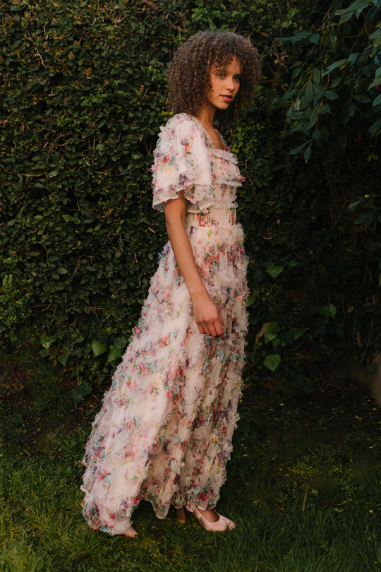 A woman with curly hair stands in a garden wearing the Ivy City Co Florentina Dress in Pink Floral—a stylish, nursing-friendly maternity dress with ruffles and short sleeves, perfect for any season and surrounded by green foliage.