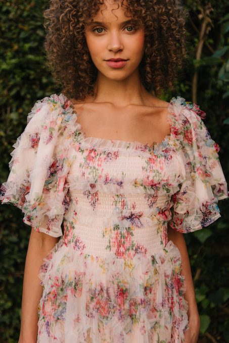 A woman with curly hair stands outdoors in the Ivy City Co Florentina Dress in Pink Floral, a nursing-friendly maternity dress with puffed sleeves, surrounded by lush greenery.