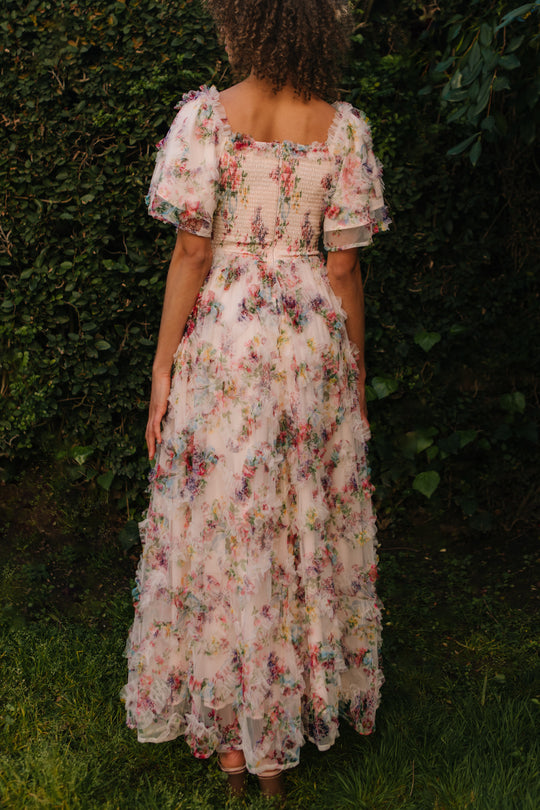 A person stands on grass, facing away, wearing the Ivy City Co Florentina Dress in Pink Floral—a pastel maternity dress with puffy short sleeves and textured fabric. Green shrubs and leafy plants are in the background.