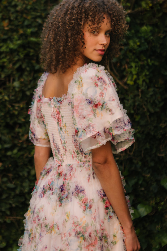 A woman with curly hair stands outdoors among green foliage, wearing the Ivy City Co Florentina Dress in Pink Floral—a light maternity dress with puffed sleeves and ruffled layers—glancing over her shoulder at the camera.