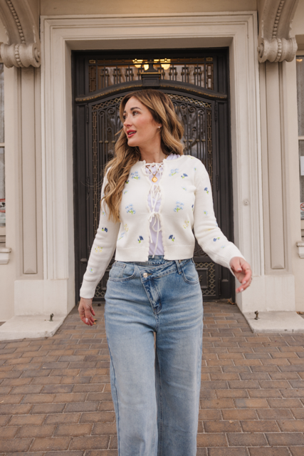 A woman with long brown hair walks on a paved path in front of an ornate building entrance, wearing the Ivy City Co Della Sweater in Cream and high-waisted blue jeans.