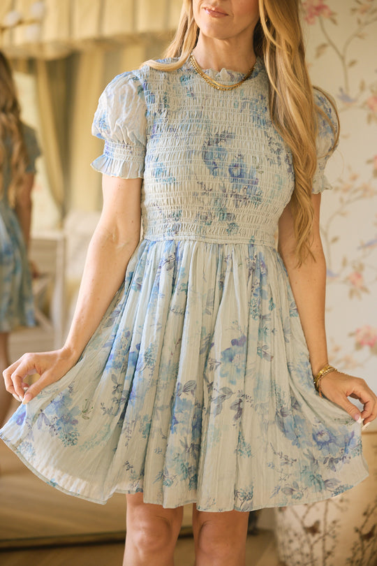 A woman in Ivy City Co's Flynn Dress in Aquarelle Fleur, a light blue watercolor floral design with a smocked bodice, stands indoors holding her skirt. She wears her long wavy blonde hair loose, accessorized with a gold necklace and bracelet.