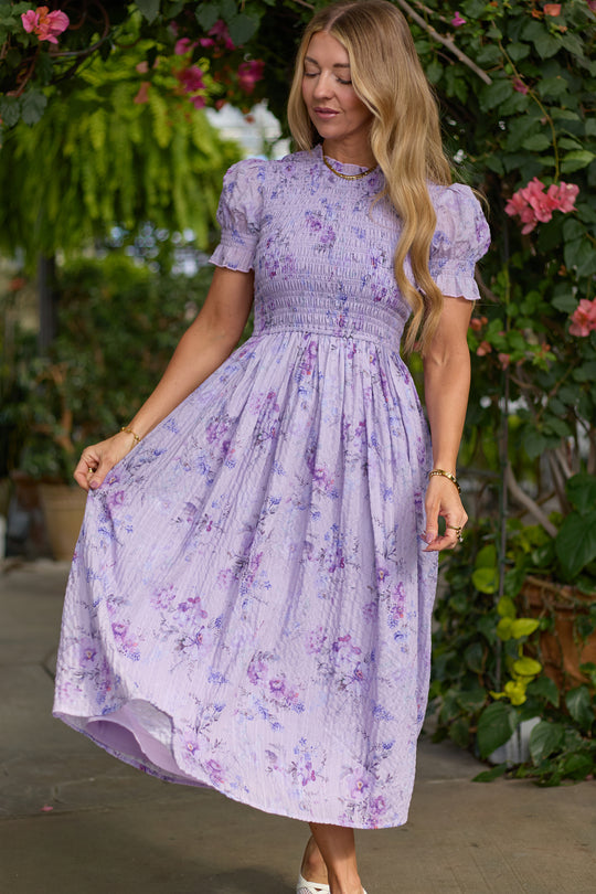 A woman stands outdoors wearing the Ivy City Co Flynn Midi Dress in Lavender Aquarelle Fleur, a floral, nursing-friendly midi with a smocked bodice. She holds out the skirt, her wavy blonde hair framed by lush greenery and pink flowers.