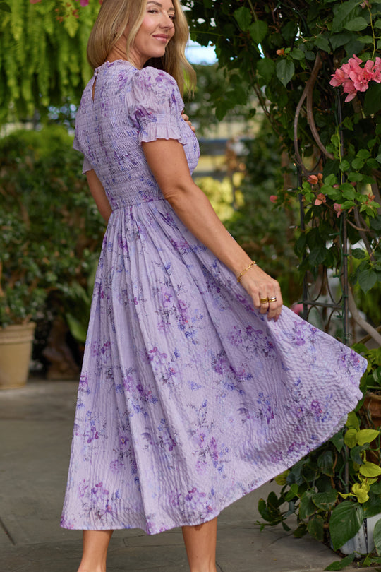 A woman wearing the Ivy City Co Flynn Midi Dress in Lavender Aquarelle Fleur stands outdoors, smiling and holding her flowing skirt, surrounded by green plants and pink flowers.