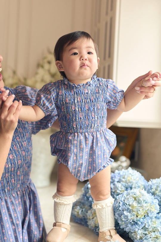 A baby in the Ivy City Co Baby Mariah Romper in Linear Print stands while holding an adult’s hand, with blue and white hydrangeas in the foreground.