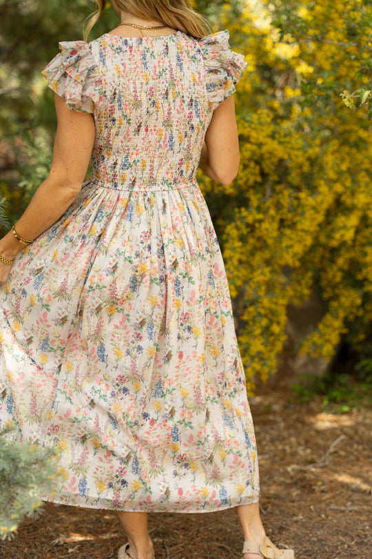 A woman stands outdoors, shown from behind, wearing the Hattie Dress in Cordelia Floral by Ivy City Co. Yellow flowers and greenery surround her in a sunlit setting, highlighting the dress’s elegant A-line midi silhouette with ruffled sleeves—a stylish and maternity-friendly choice.