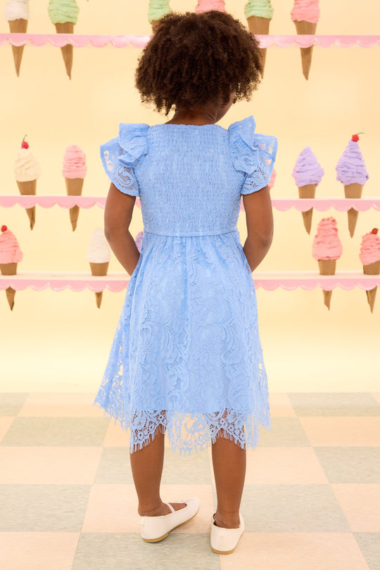 A young girl wearing the Mini Hattie Dress in Cornflower Blue Lace by Ivy City Co and white shoes stands with her back to the camera in front of a pastel wall decorated with colorful ice cream cones.