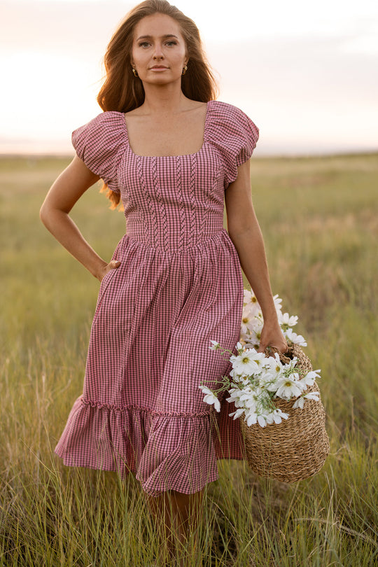 A woman wearing the Monica Dress in Pink & Brown Gingham by Ivy City Co stands in a grassy field at sunset, holding a woven basket filled with white flowers.
