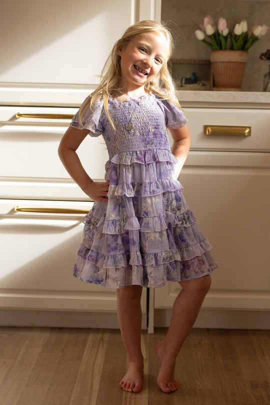 A smiling young girl with long blonde hair stands barefoot in a kitchen, wearing the Ivy City Co Mini Winnie Dress in Lavender Aquarelle Fleur. Sunlight streams in as a vase of tulips sits on the counter in the background.