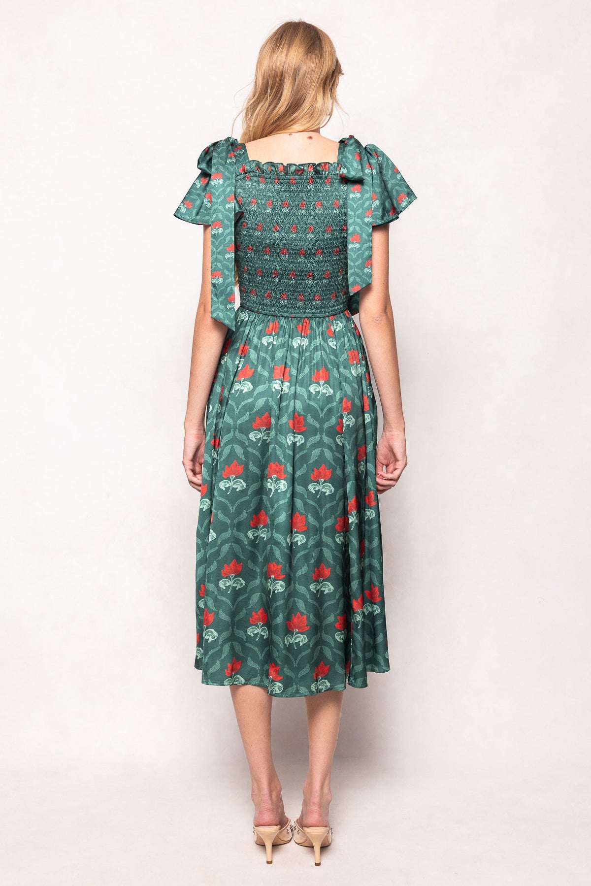 A woman stands facing away in the Ivy City Co Rayna Dress in Green Floral Block, featuring red patterns, ruffled short sleeves, and a smocked bodice that’s both nursing and maternity friendly. She pairs it with beige heels against a plain light background.