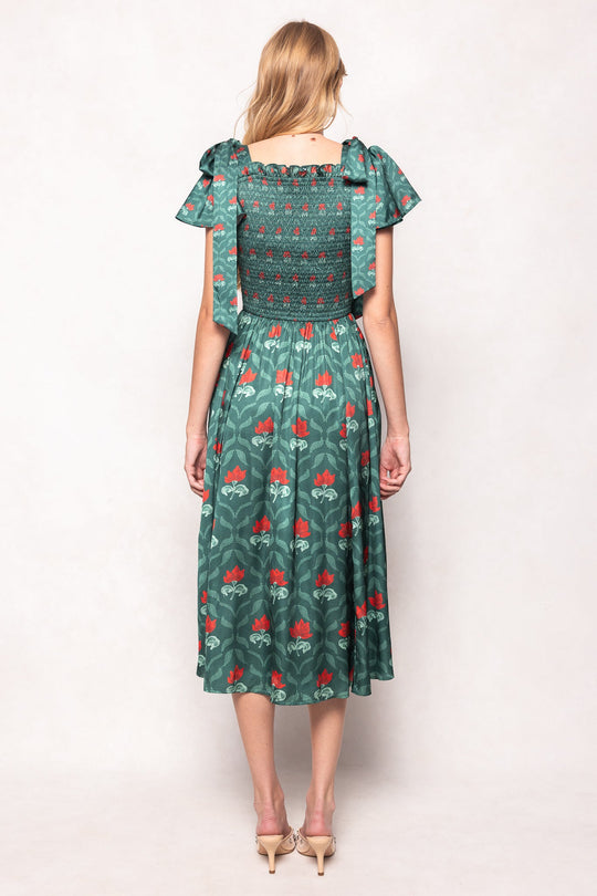 A woman stands facing away in the Ivy City Co Rayna Dress in Green Floral Block, featuring red patterns, ruffled short sleeves, and a smocked bodice that’s both nursing and maternity friendly. She pairs it with beige heels against a plain light background.