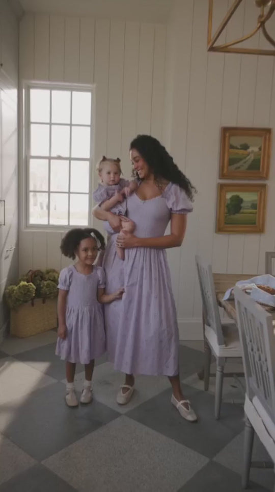 A woman and two young girls, all dressed in matching Ivy City Co Emmi Dresses in Lilac, stand smiling together in a bright dining room accented by large windows and framed artwork. Their beautifully coordinated outfits are not only stylish but also maternity-friendly, ensuring comfort for every occasion.