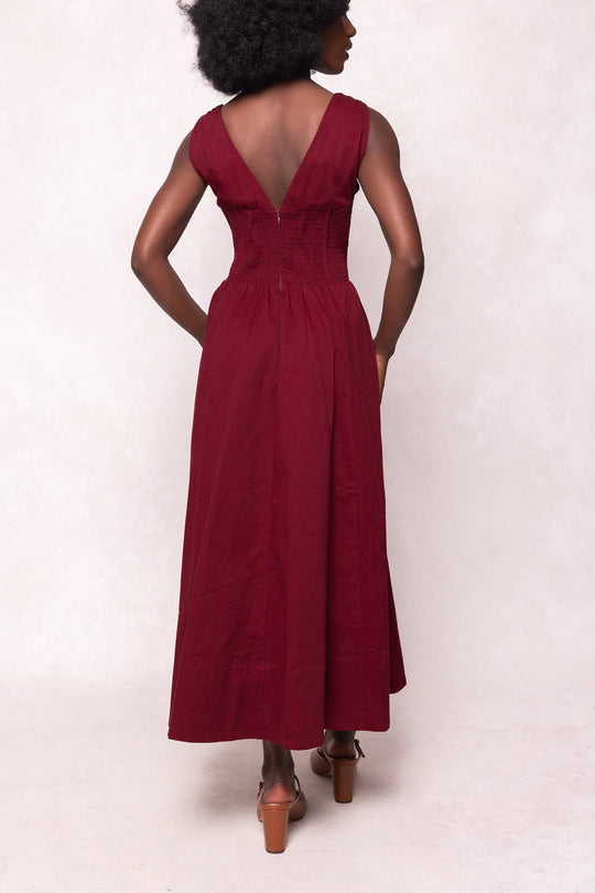 A woman stands facing away, wearing the Ivy City Co Kayla Dress in Burgundy. The sleeveless midi dress features a V-shaped back. She has curly hair and brown heeled sandals, with one hand resting on her hip.