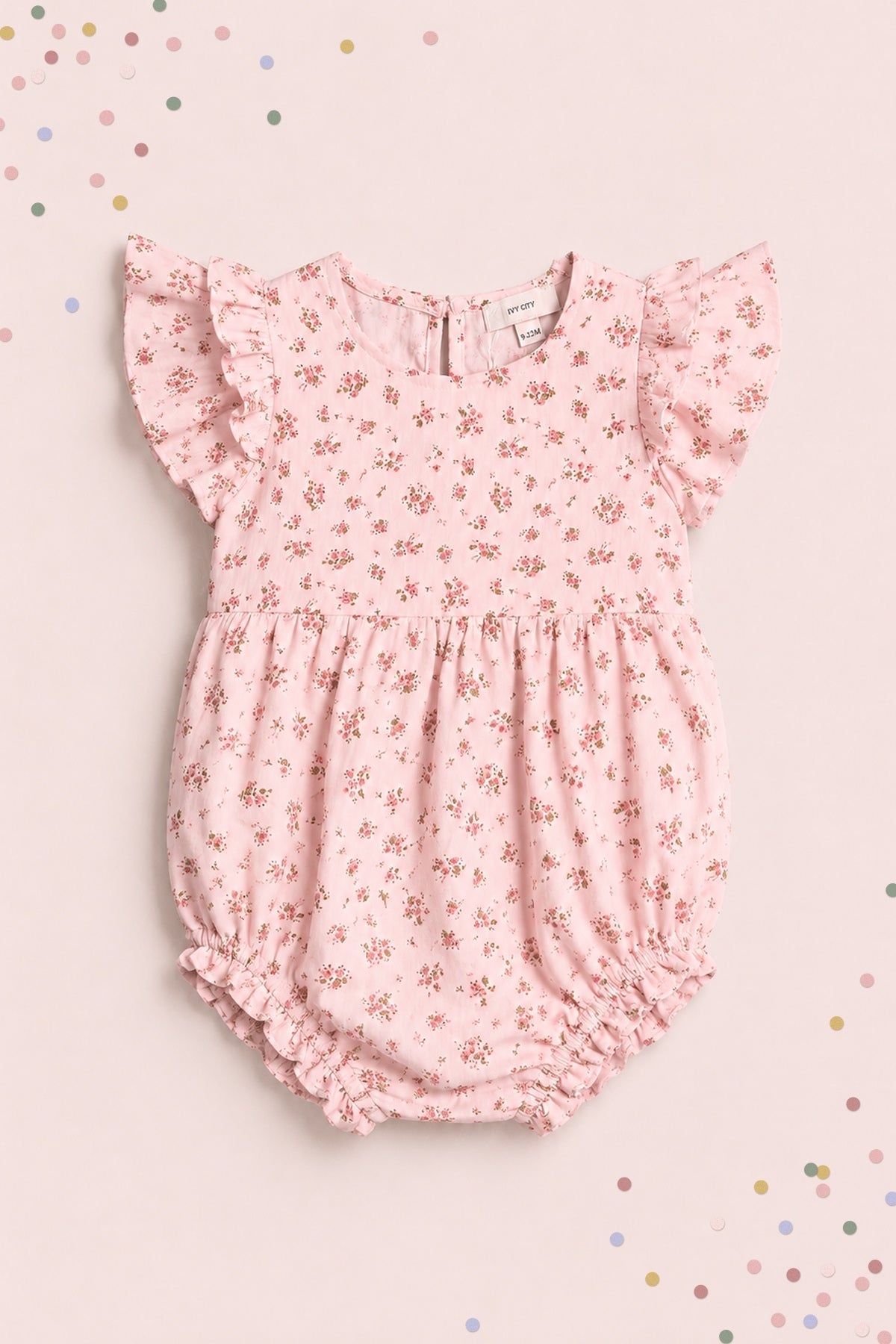 The Ivy City Co Baby Hattie Romper in Madeline Pink, featuring flutter sleeves and a delicate floral pattern, is pictured laid flat on a light pink background sprinkled with pastel confetti—an adorable choice for a first birthday outfit.