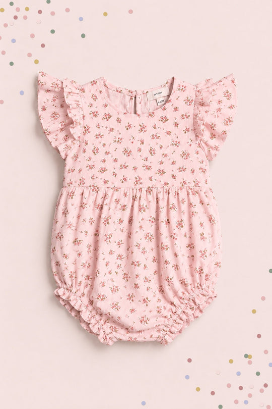 The Ivy City Co Baby Hattie Romper in Madeline Pink, featuring flutter sleeves and a delicate floral pattern, is pictured laid flat on a light pink background sprinkled with pastel confetti—an adorable choice for a first birthday outfit.