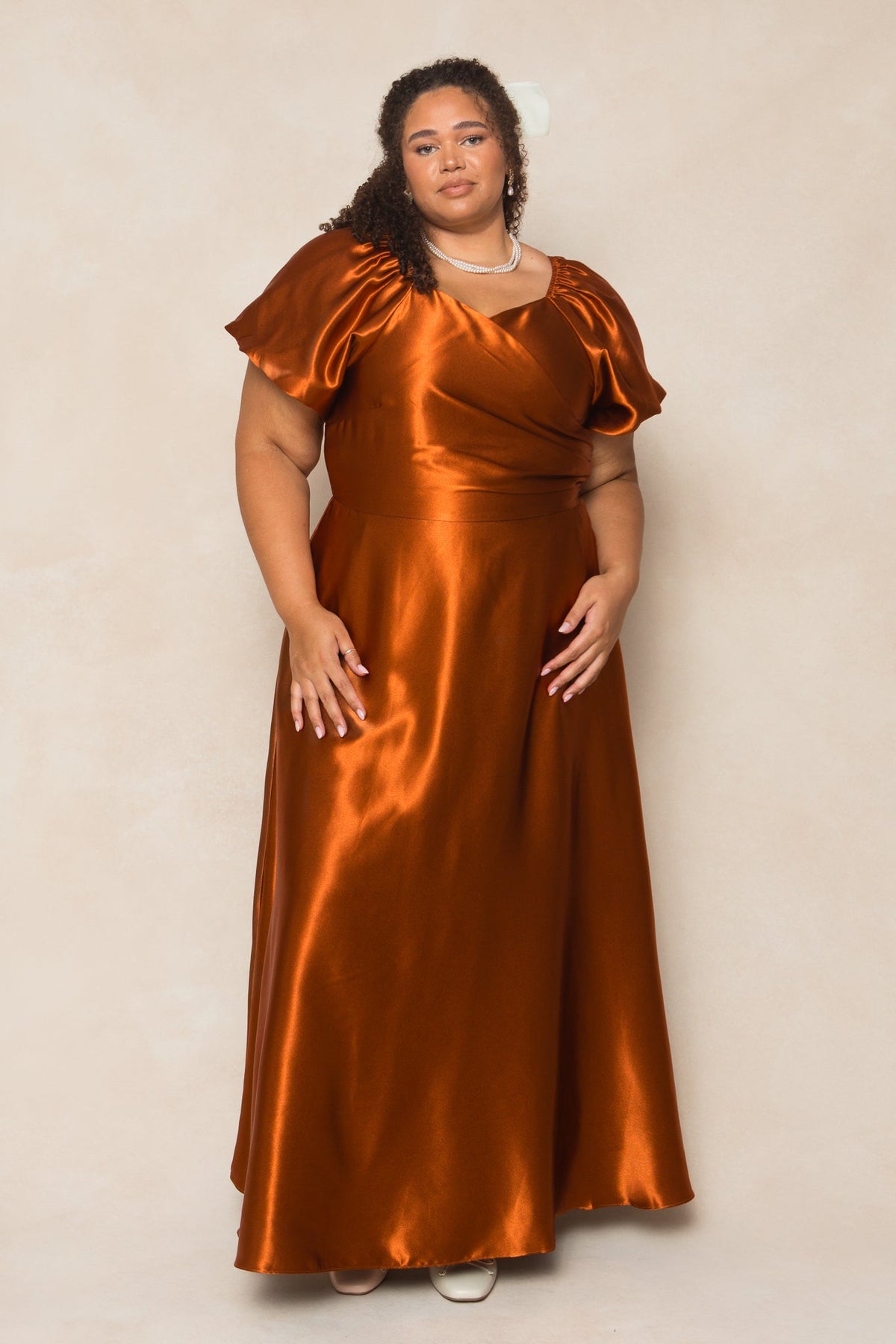 A woman with curly hair, accessorized with a necklace, stands against a light beige background wearing the Ivy City Co Tessie Dress in Amber Satin—a shiny, floor-length gown with short puff sleeves—making it an ideal choice for a fall bridesmaid’s look.