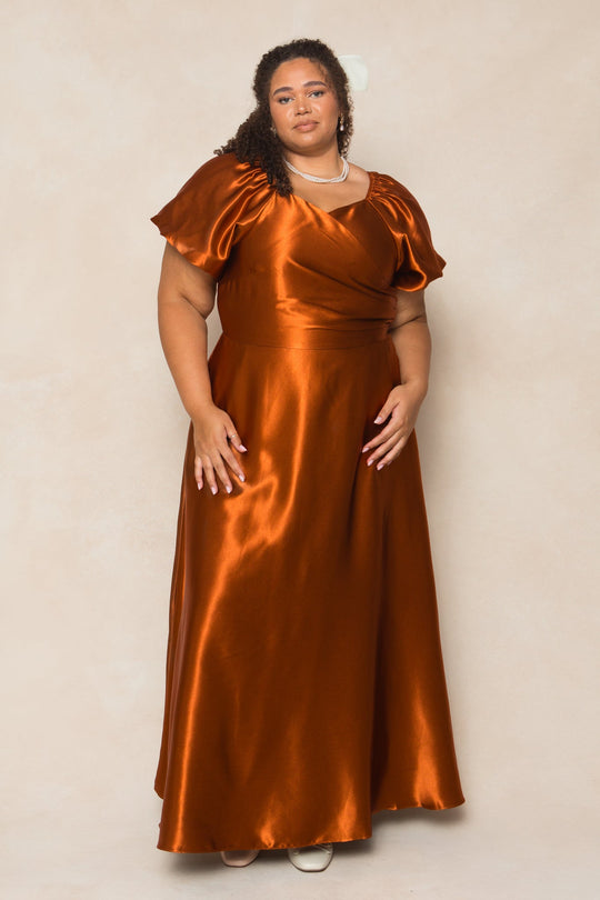 A woman with curly hair, accessorized with a necklace, stands against a light beige background wearing the Ivy City Co Tessie Dress in Amber Satin—a shiny, floor-length gown with short puff sleeves—making it an ideal choice for a fall bridesmaid’s look.