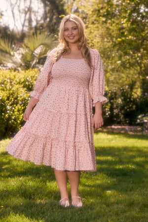 A woman stands on grass in a sunlit garden, wearing Ivy City Co’s Madeline Dress In Pink—a maternity-friendly, pink, long-sleeved tiered dress with a small floral pattern and light pink flats. Trees and bushes are in the background.