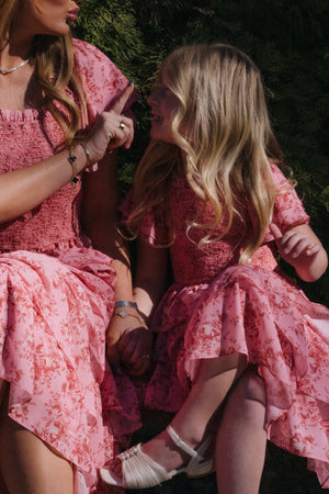 A woman and a young girl, both wearing matching Ivy City Co Mini Grace Dresses in Desert Heritage Rose, sit close together outdoors, holding hands and looking at each other. The woman gestures with her finger while the girl listens attentively.