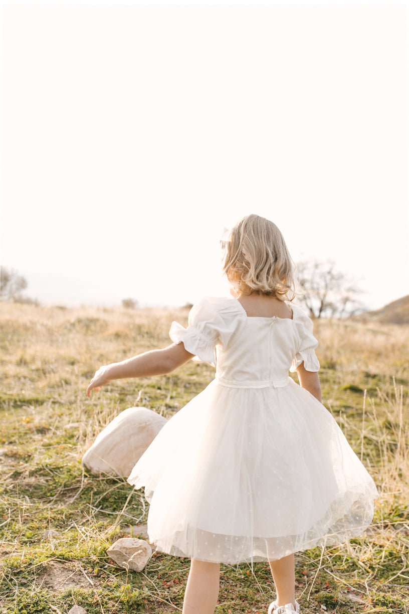 A young child wearing the Ivy City Co Mini Ballerina Dress in White stands in a grassy field with their back to the camera, reaching toward a rock beneath a bright, clear sky.