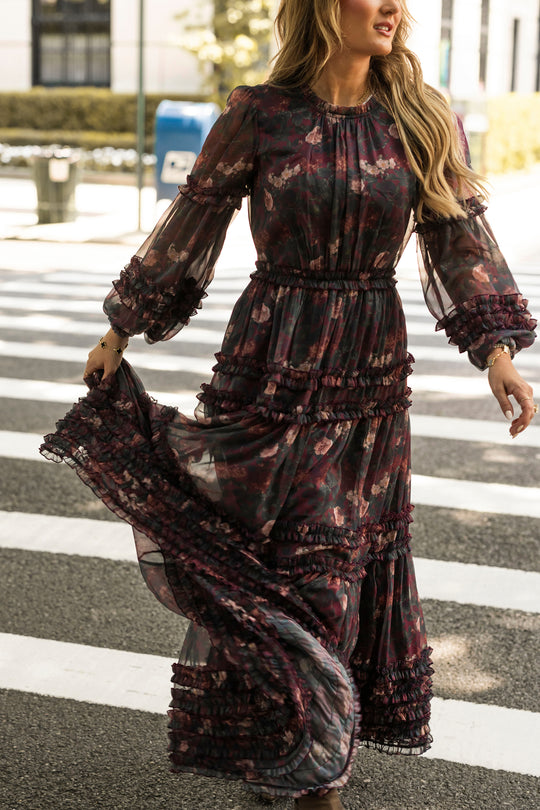 A woman in the Geneva Dress in Aubergine Resplendent Bloom by Ivy City Co—a dark floral, long-sleeved, ruffled maxi dress perfect for fall weddings—crosses a street while holding up part of her skirt. In the background are a blue mailbox and blurred buildings.