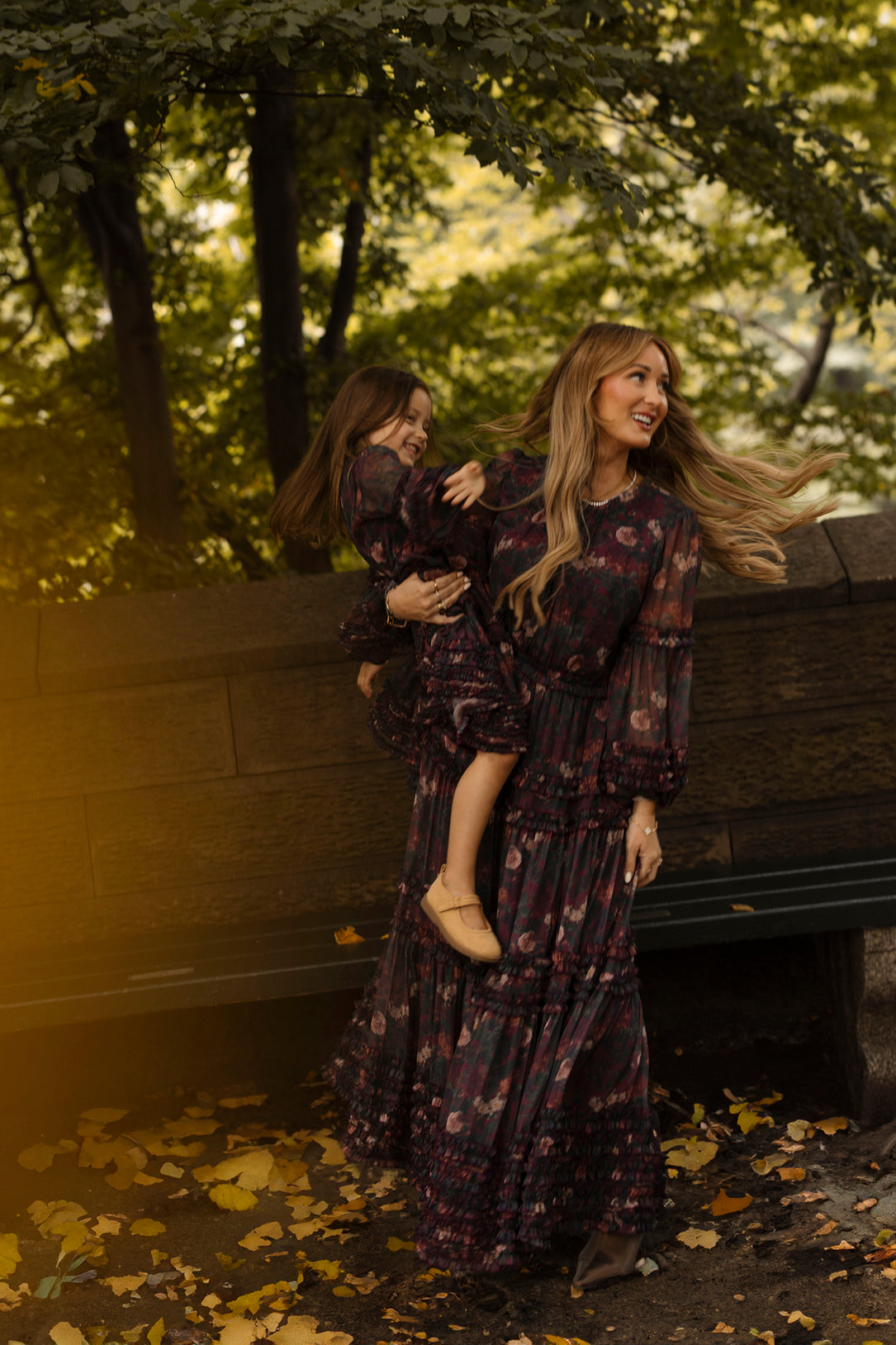A woman wearing the Ivy City Co Geneva Dress in Aubergine Resplendent Bloom holds a smiling young girl, also dressed in matching Ivy City Co attire, outdoors beside a bench and surrounded by trees with fallen yellow leaves.