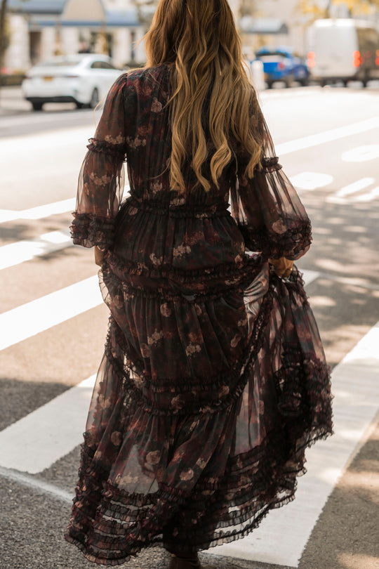 A person with long wavy hair wearing the Geneva Dress in Aubergine Resplendent Bloom by Ivy City Co crosses a street at a crosswalk on a sunny day.