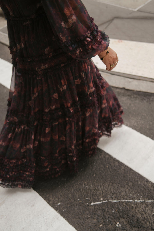 A person wearing the Ivy City Co Geneva Dress in Aubergine Resplendent Bloom crosses the street, shown from the waist down. Their hand, adorned with rings, is visible near a crosswalk and sidewalk.