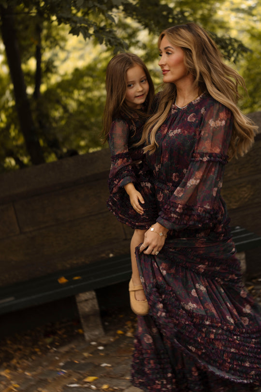 A woman wearing the Geneva Dress in Aubergine Resplendent Bloom by Ivy City Co carries a young girl dressed in a matching balloon sleeve dress. They are outdoors near a wooden bench, surrounded by trees and greenery.