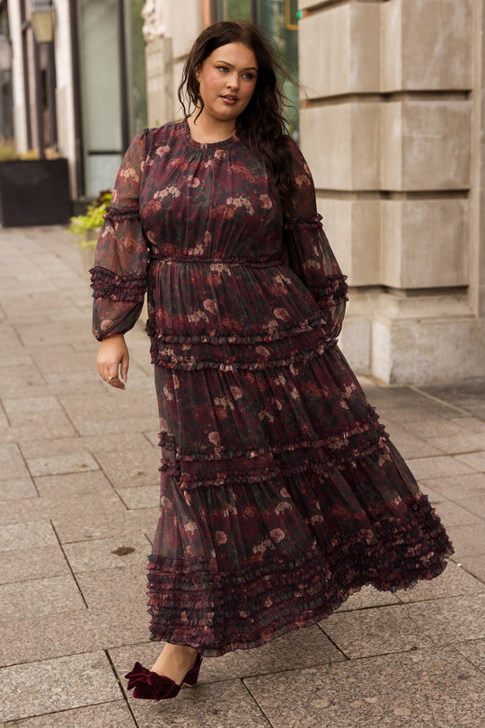 A woman wearing the Ivy City Co Geneva Dress in Aubergine Resplendent Bloom, featuring elegant balloon sleeves, pairs it with red shoes as she walks along a city sidewalk near a stone building, glancing slightly to her left.