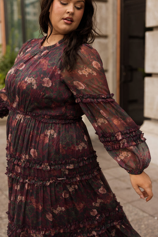 A woman wearing the Geneva Dress in Aubergine Resplendent Bloom by Ivy City Co stands outdoors, gently swaying with one arm extended. Her long dark hair cascades down as she looks downward, surrounded by modern buildings.