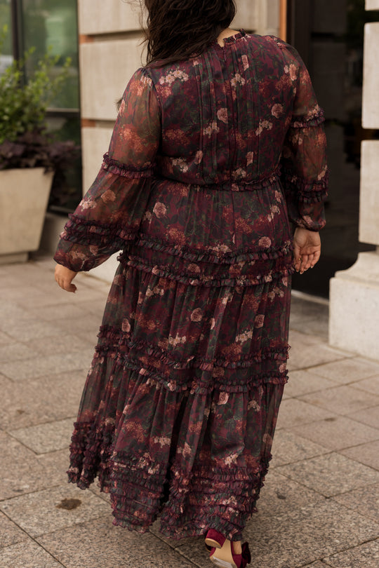 Wearing the Geneva Dress in Aubergine Resplendent Bloom by Ivy City Co—a long, sheer, dark floral fall maxi dress with ruffle details—a person walks on a stone sidewalk, facing away from the camera.