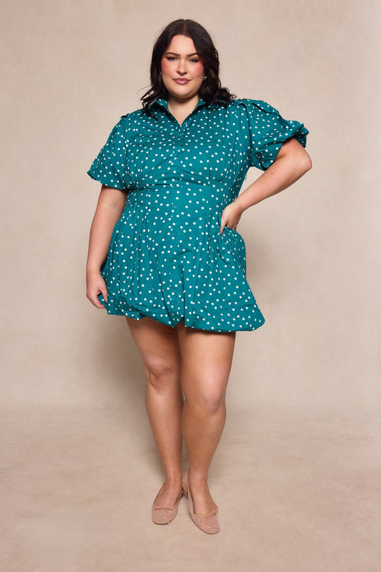 A woman wearing the Ivy City Co Cody Dress in Deep Teal Polka Dot stands confidently with one hand on her hip and the other in her pocket, posing against a neutral background.