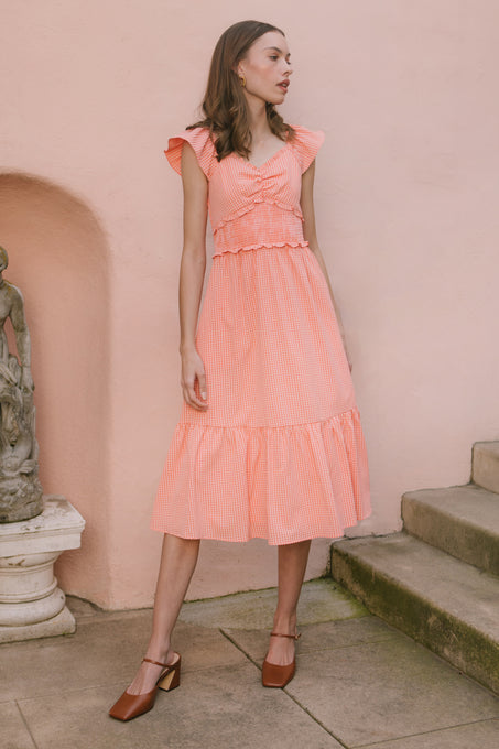 A woman in the Ivy City Co Gidget Dress in Orange Gingham and brown heels stands by a stone statue and stairs, with a light pink wall behind her.