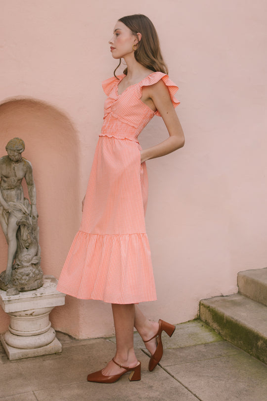 A woman in the Ivy City Co Gidget Dress in Orange Gingham with ruffled sleeves stands by a stone statue and pink wall, looking to the side. She pairs the maternity dress with brown heels and wears her long, wavy hair down.