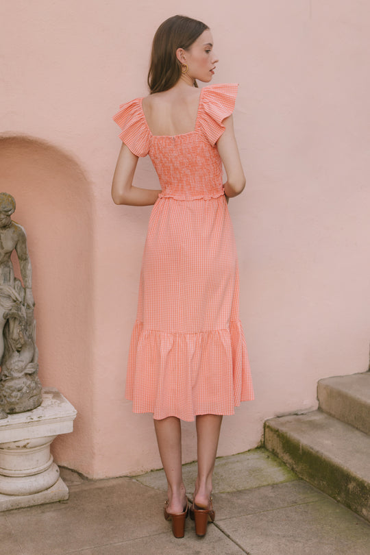 A woman in the Ivy City Co Gidget Dress in Orange Gingham, featuring ruffled sleeves and a smocked bodice, stands on a stone path near a pale pink wall and a small statue, facing away from the camera.