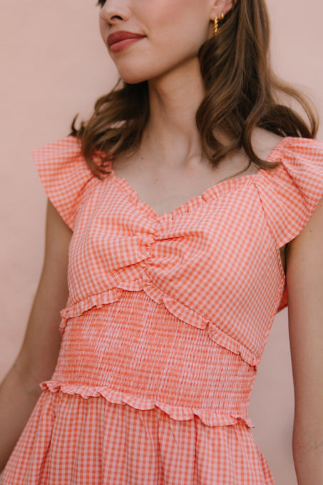 A woman with wavy hair and gold hoop earrings wears the Ivy City Co Gidget Dress in orange gingham, featuring ruffled sleeves and a ruched bodice, standing against a light pink background.