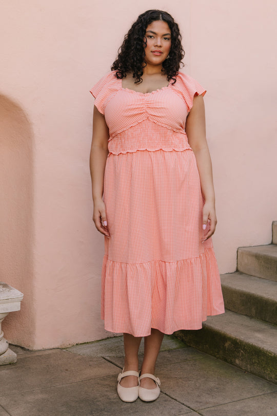 A woman with curly hair wearing the Ivy City Co Gidget Dress in Orange Gingham and beige shoes stands by stone steps against a pale pink wall.