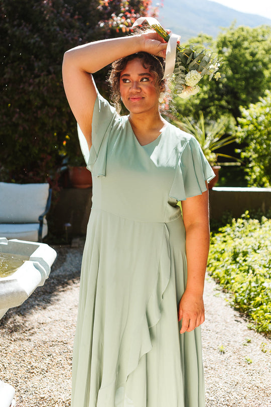 A woman stands outdoors surrounded by greenery, holding a bouquet above her head while sunlight illuminates her face. She is wearing the Callie Dress in Sage Chiffon by Ivy City Co.