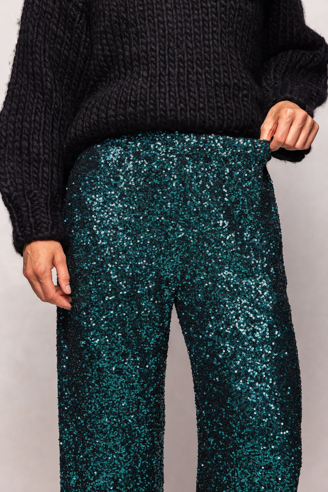 Person wearing dark green sequin pants with a black sweater on a light gray background