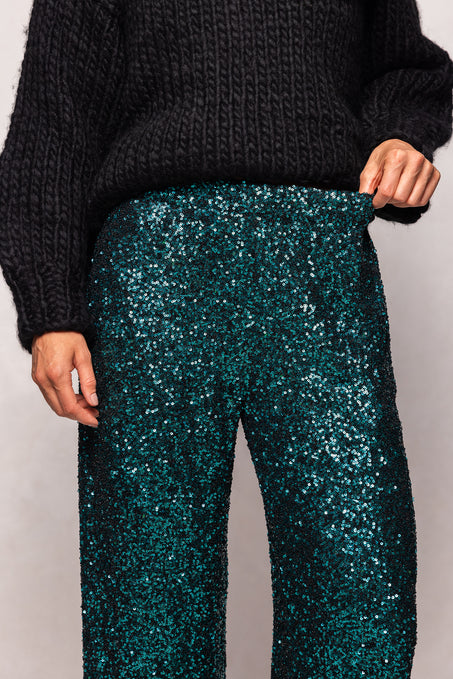 Person wearing dark green sequin pants with a black sweater on a light gray background