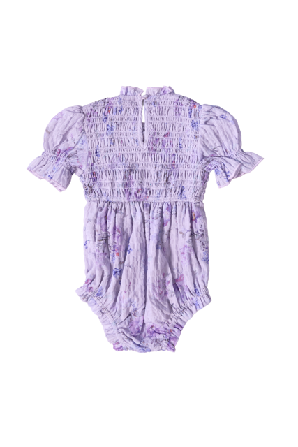 The Ivy City Co Baby Flynn Romper in Lavender Aquarelle Fleur features a soft, textured fabric with a subtle watercolor floral print, puffed short sleeves, a smocked bodice, and back button details. Displayed on a white background.