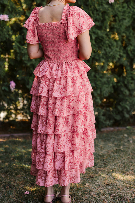 A person wearing the Ivy City Co Grace Dress in Desert Heritage Rose stands on grass outdoors, facing away from the camera with green trees in the background.