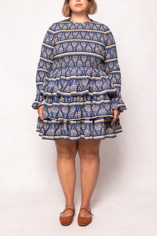 Someone wearing the Greer Dress in Navy Fleur by Ivy City Co, featuring a smocked bodice and tiered skirt, paired with brown flats, stands against a plain light background.